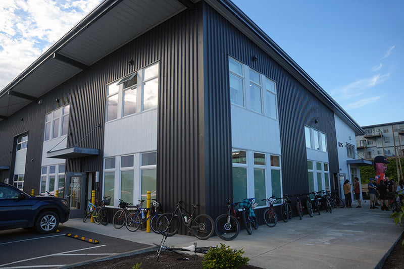 A modern two-story building with dark gray vertical siding and white paneling, with several bicycles parked along the sidewalk in front. A blue car is partially visible on the left, and a few people are standing near the right side of the building under a blue sky.
