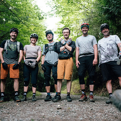 Six mountain biker friends wearing helmets, knee pads, and shorts stand together on a dirt path in a wooded forest setting.