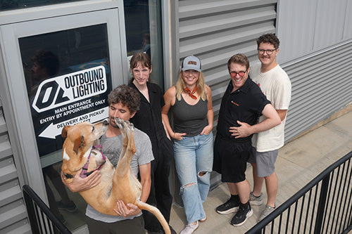Five people and a large tan dog pose together on the front steps of a building with a gray corrugated metal exterior. One person holds the dog upside down.