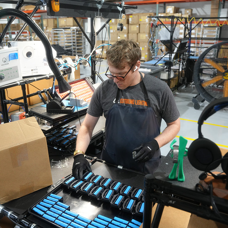A worker in a warehouse setting wearing a dark gray t-shirt and a navy blue apron with orange text that reads 'OUTBOUND LIGHTING'. The person is wearing black gloves and earplugs while organizing blue and black battery packs in a tray on a workbench.