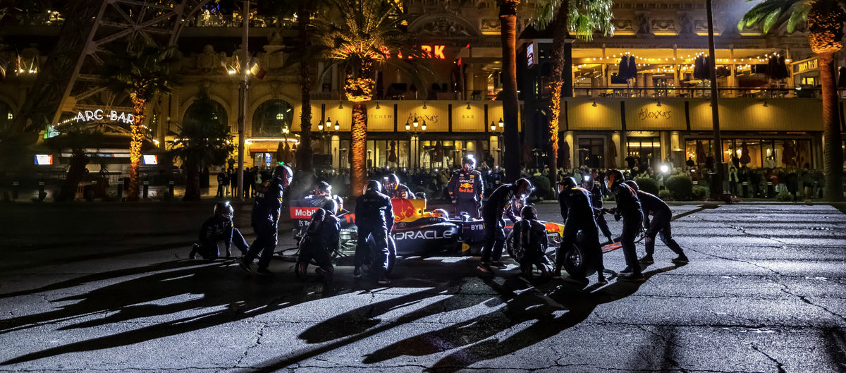 A race team in black uniforms works around a red and black Formula 1 car at night on a Las Vegas Strip street, with the Eiffel Tower and brightly lit hotels in the background.