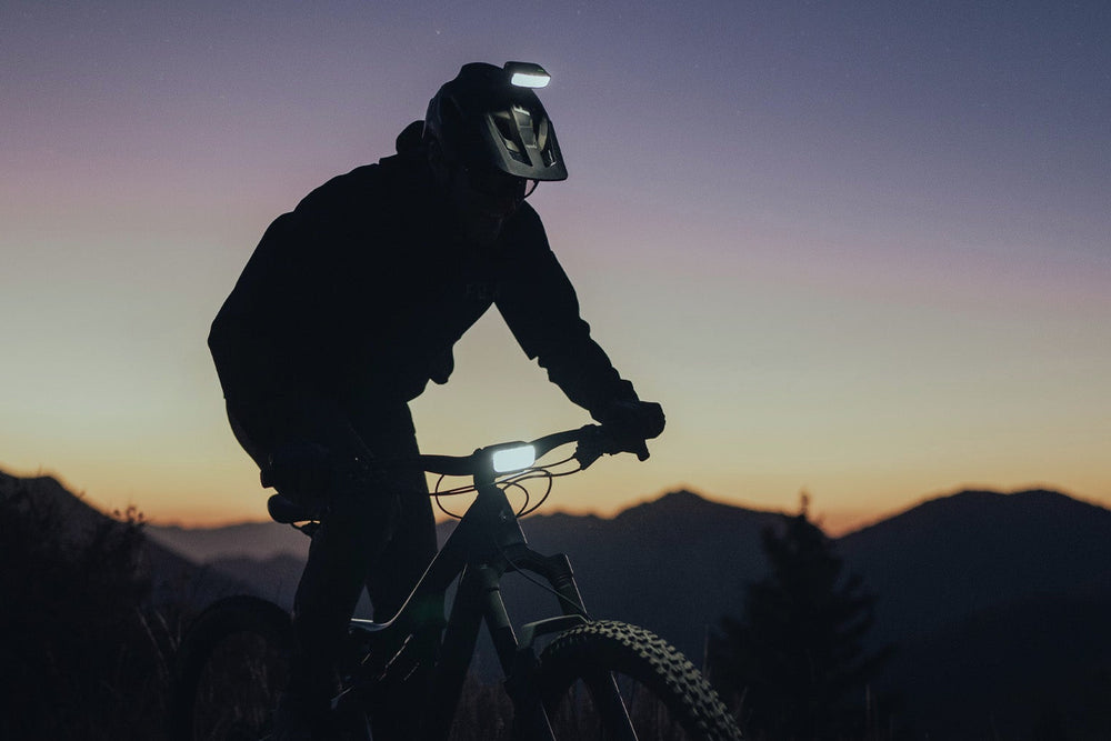 A mountain biker in silhouette riding at dusk with a twilight sky and distant mountains in the background.